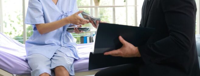 A woman sitting on a hospital bed receives documents from a man in a black suit.