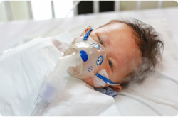 A baby lies on a hospital bed wearing an oxygen mask.